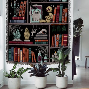 Op de afbeelding: Een zwart-witte wandkleed met een boekenkastontwerp. De planken zijn gevuld met boeken, kaarsen, kristallen en andere heksachtige items. De tekst "OUIJA" is zichtbaar op een van de planken.