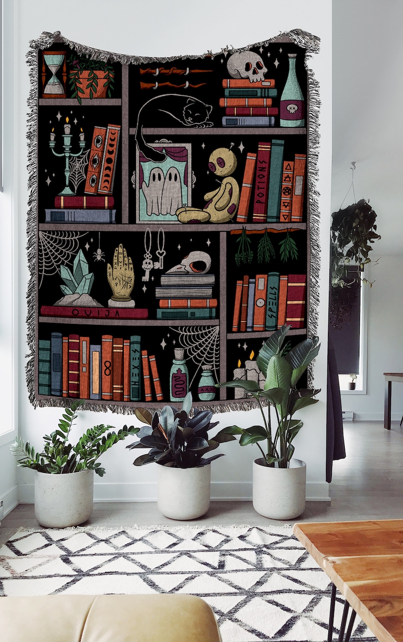 Op de afbeelding: Een zwart-witte wandkleed met een boekenkastontwerp. De planken zijn gevuld met boeken, kaarsen, kristallen en andere heksachtige items. De tekst "OUIJA" is zichtbaar op een van de planken.