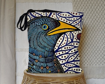Könnte beinhalten: Eine blaue und gelbe Tragetasche mit einem farbenfrohen Vogeldesign. Der Vogel hat einen blauen Kopf, einen gelben Schnabel und rote und weiße Akzente. Der Hintergrund ist weiß mit blauen und roten Blattmustern.