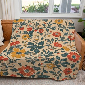May include: A floral patterned throw blanket draped over a brown leather couch. The blanket features a cream background with a repeating pattern of red, yellow, and teal flowers and leaves. A white pillow is on the couch.