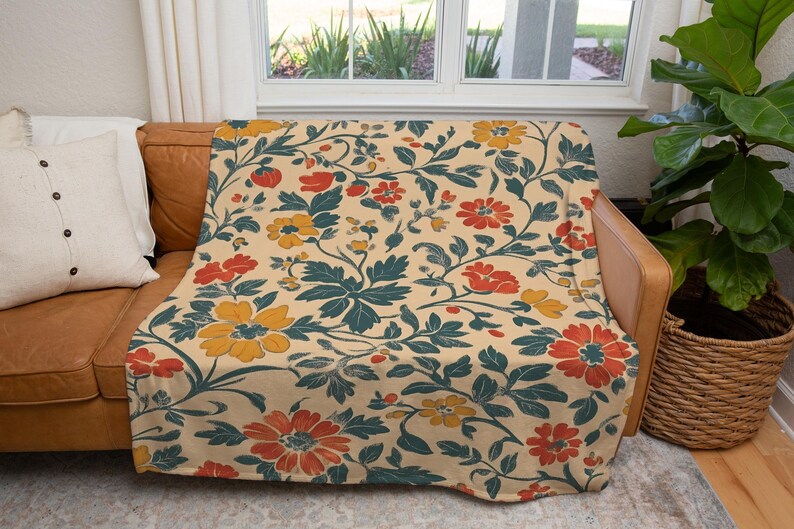 May include: A floral patterned throw blanket draped over a brown leather couch. The blanket features a cream background with a repeating pattern of red, yellow, and teal flowers and leaves. A white pillow is on the couch.