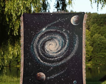 Può includere: Un arazzo tessuto nero con un design a galassia che presenta una spirale vorticosa di stelle e pianeti. L'arazzo è frangiato su tutti i lati ed è tenuto da una persona davanti a un campo erboso verde.