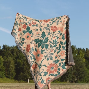 May include: A beige floral throw blanket with a repeating pattern of red and peach flowers and green leaves. The blanket has a fringed edge and is held up against a blue sky background.