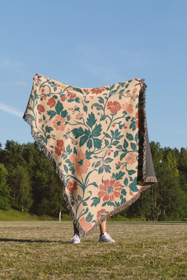 May include: A beige floral throw blanket with a repeating pattern of red and peach flowers and green leaves. The blanket has a fringed edge and is held up against a blue sky background.