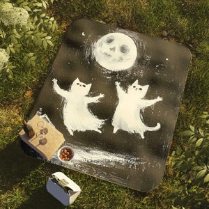 May include: Black and white picnic blanket with a moon and two cats painted on it. The cats are white and have a whimsical, cartoon-like style.
