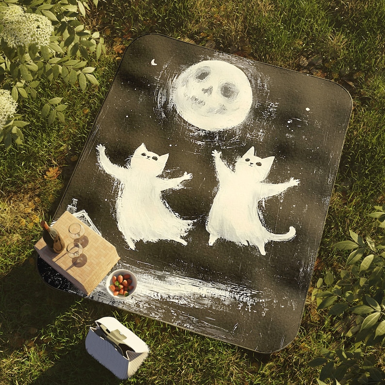 May include: Black and white picnic blanket with a moon and two cats painted on it. The cats are white and have a whimsical, cartoon-like style.