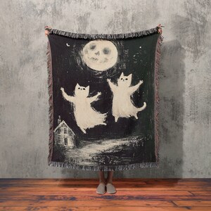 May include: A black woven throw blanket with a white moon and two white cats dancing in front of a small white house. The blanket has fringe around the edges.