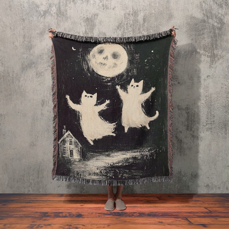 May include: A black woven throw blanket with a white moon and two white cats dancing in front of a small white house. The blanket has fringe around the edges.