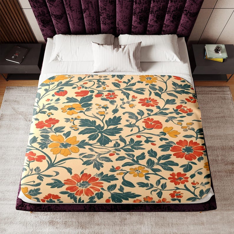 May include: A floral patterned blanket featuring a cream background with orange and yellow flowers, and teal green leaves. The blanket is on a bed with white pillows and a dark purple headboard.
