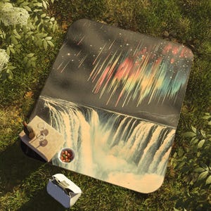 May include: A picnic blanket with a colorful aurora borealis and waterfall design. The blanket is laid out on green grass with a wine bottle, glasses, and a bowl of nuts on top.