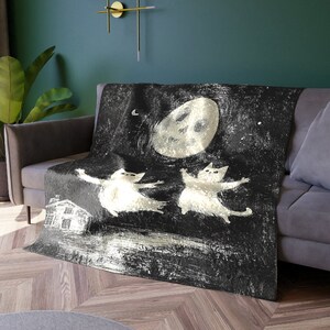 May include: A black and white throw blanket with a whimsical design of two cats dancing in front of a large moon. The background is a dark starry night.