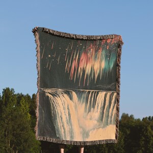 May include: A woven tapestry throw blanket with a dark background featuring a colorful aurora borealis above a waterfall. The blanket has a fringe border.