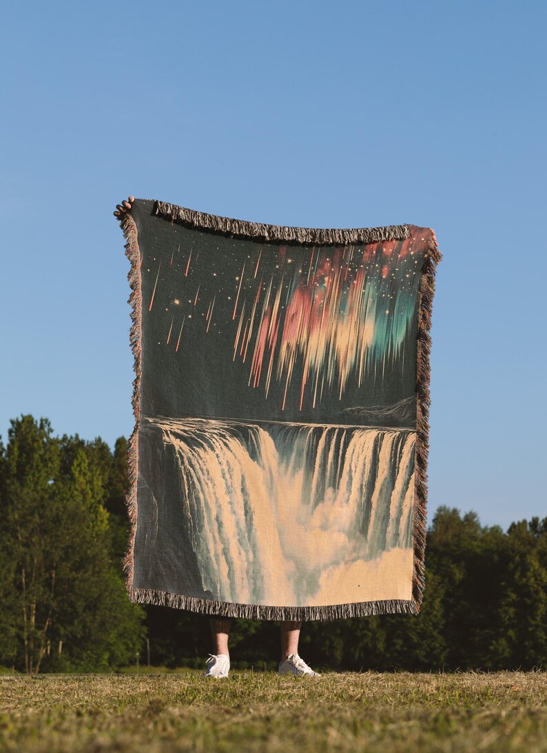 May include: A woven tapestry throw blanket with a dark background featuring a colorful aurora borealis above a waterfall. The blanket has a fringe border.