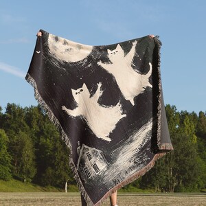 May include: A black and white woven tapestry featuring three white cats flying in front of a full moon and a house. The tapestry is being held up by a person in a grassy field.