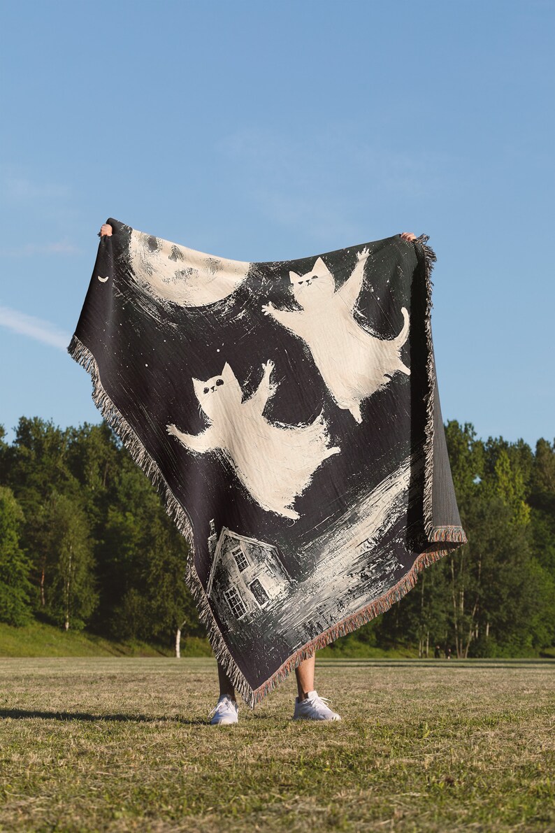 May include: A black and white woven tapestry featuring three white cats flying in front of a full moon and a house. The tapestry is being held up by a person in a grassy field.