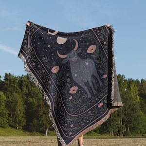 Könnte beinhalten: Eine dunkelblaue Decke mit einem Sternbild-Design, das eine Stier-Illustration, Halbmonde, Sterne und florale Details zeigt. Die Decke hat einen Fransenrand und wird vor blauem Himmel und grünen Bäumen gehalten.