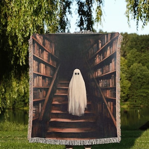 May include: A woven tapestry throw blanket features a spooky image of a ghost standing on a staircase in a library. The ghost is white with two black eyes. The library has bookshelves on either side of the staircase.
