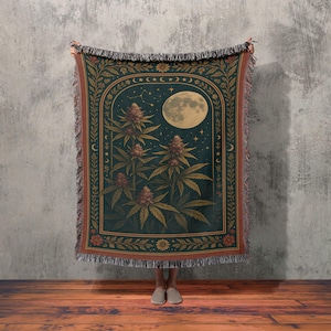 May include: A woven throw blanket featuring a dark teal background with a full moon, stars, and cannabis plants. The design is framed by an archway with floral and celestial elements. The blanket has a fringed edge.