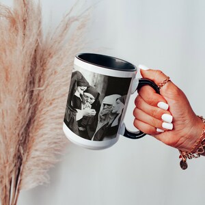May include: White ceramic mug with a black rim and handle. The mug features a black and white photograph of three people in a group, smoking cigarettes.