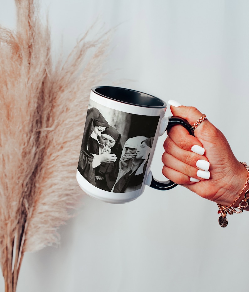 May include: White ceramic mug with a black rim and handle. The mug features a black and white photograph of three people in a group, smoking cigarettes.