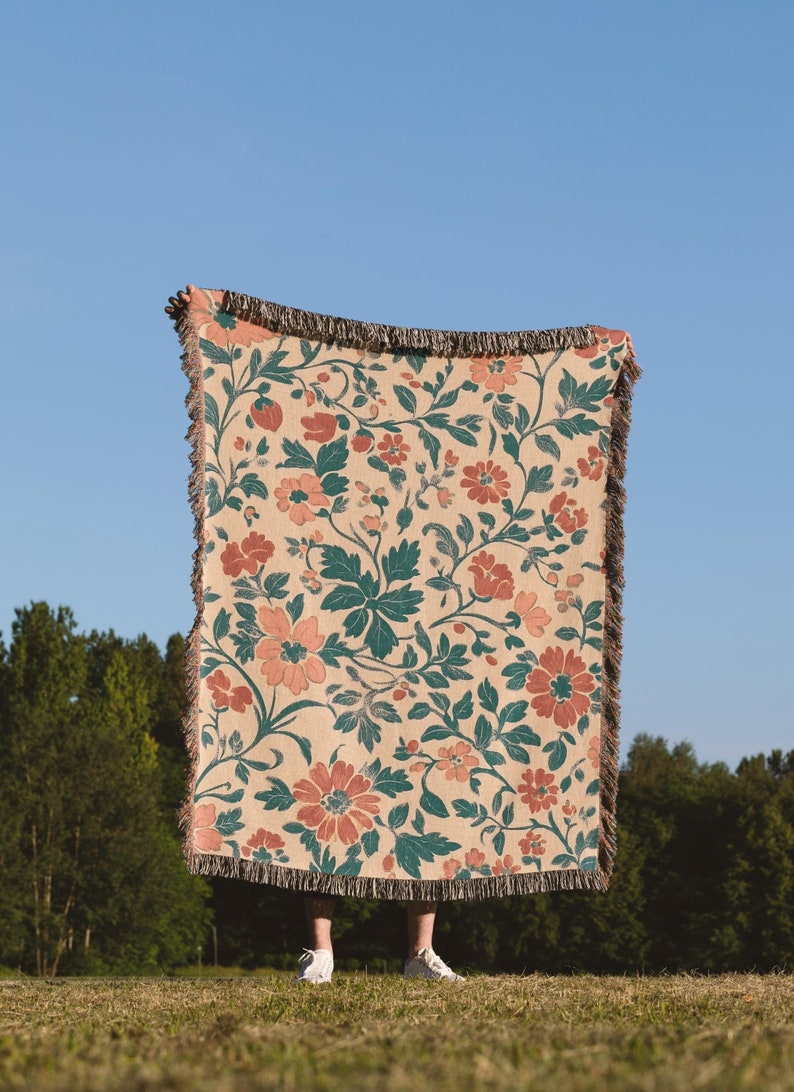 May include: A beige floral throw blanket with a repeating pattern of coral-colored flowers and teal leaves. The blanket has a fringed edge and is held up against a clear blue sky. The blanket is made of a woven material.