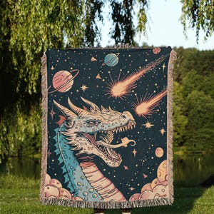 May include: A woven tapestry featuring a blue and pink dragon with a starry night sky background. The dragon is facing the viewer with its mouth open and has a comet shooting past its head. The tapestry has a fringe border.