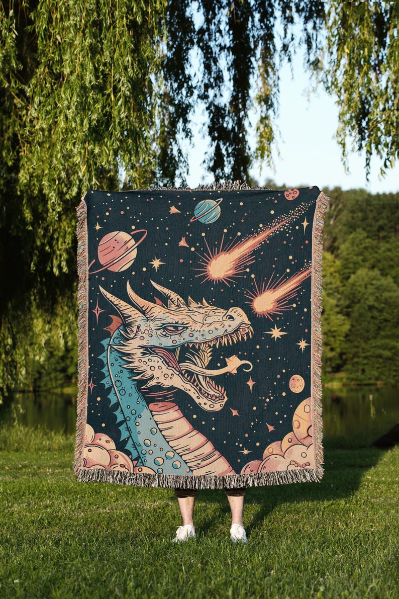 May include: A woven tapestry featuring a blue and pink dragon with a starry night sky background. The dragon is facing the viewer with its mouth open and has a comet shooting past its head. The tapestry has a fringe border.