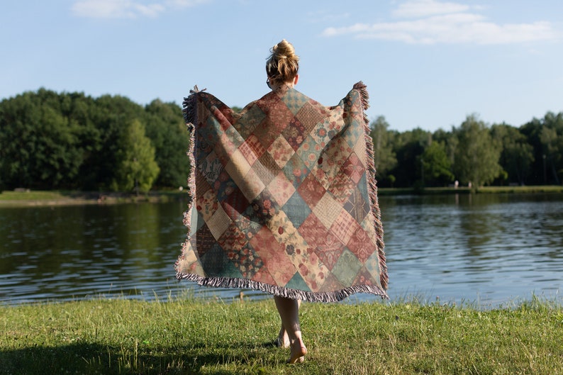 May include: A person standing by a lake wearing a colourful patchwork throw blanket with fringe.