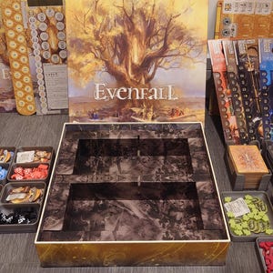 May include: A game organiser for the board game Evenfall. The organiser is made of cardboard and has a brown and black design. The organiser has two compartments for storing game pieces. The organiser is sitting on a table with other game components.