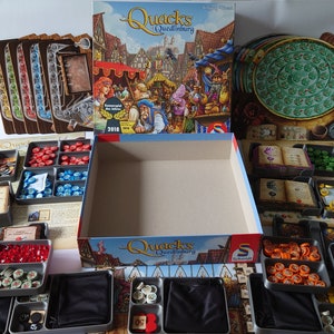 May include: A game box with the title "Quacks of Quedlinburg" and various game components, including a game board, cards, tokens, and dice. The box is open and the components are organised in trays.