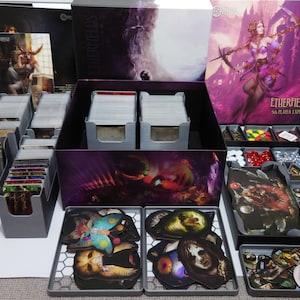 May include: A grey and black organiser box with compartments for storing game pieces and cards. The box has a purple and black design with a woman on the front. The text on the box reads "Etherfields Six Player Expansion".