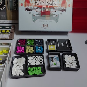 May include: A game board with a box and various components, including a game board, a box, and several trays of game pieces. The game pieces are in different colours, including red, green, blue, and white. The box has the title "Kanban EV" on it.