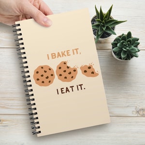 May include: A spiral-bound notebook with a light beige cover featuring the text I BAKE IT, I EAT IT in brown letters. The design includes three chocolate chip cookie illustrations, with the middle cookie showing a bite mark and the third being a fragment.