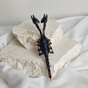 May include: A black scorpion ornament with prominent pincers and a segmented body. The scorpion is displayed on a light-coloured, textured stone surface, against a white fabric backdrop. The scorpion's tail curves upwards, ending in a stinger.