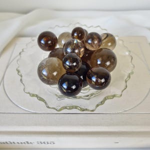 May include: A collection of smoky quartz crystal spheres in various sizes, displayed in a clear glass bowl with a scalloped edge.