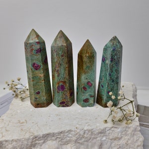 May include: Four green and pink ruby zoisite crystal points on a white stone surface. The points are all roughly the same size and shape, and they have a natural, rough texture.