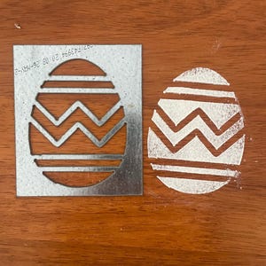 May include: A metal stencil of an egg with a zig-zag and striped pattern. The stencil is placed next to a wooden surface with a white painted egg using the stencil.