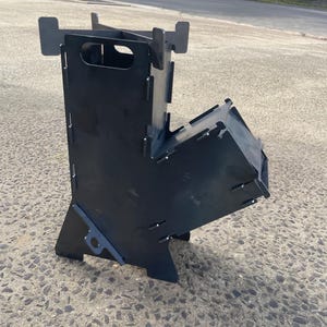 May include: A black metal wood stove with a rectangular body and angled sides. The stove has slots and tabs for assembly and ventilation. The design includes a top opening and a lower angled section for fuel.