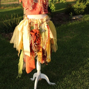 May include: A red and yellow layered skirt with a white top and a brown pendant. The skirt is made of various fabrics and has a layered, flowing design.