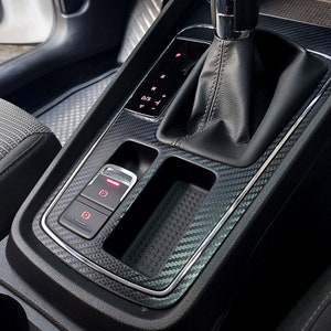 Puede incluir: Primer plano del interior de un coche negro con un patr&oacute;n de fibra de carbono. La imagen muestra una palanca de cambios, un compartimento de almacenamiento y un panel de botones con una luz roja. La palanca de cambios tiene una funda de cuero negro y una perilla de cambio de marchas plateada. El compartimento de almacenamiento est&aacute; vac&iacute;o y tiene una alfombra de goma negra. El panel de botones tiene tres botones con iconos para el freno de estacionamiento, las luces de emergencia y un tercer bot&oacute;n con un icono poco claro. La palanca de cambios tiene una pantalla que muestra las letras "P", "R", "N", "D", "S" y "-".