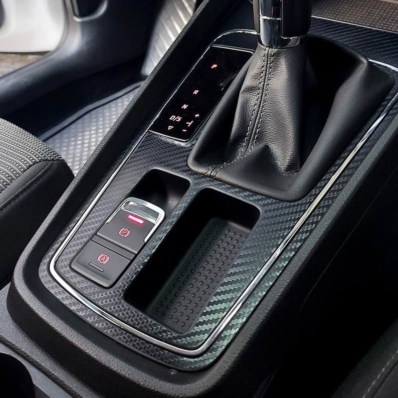 Puede incluir: Primer plano del interior de un coche negro con un patr&oacute;n de fibra de carbono. La imagen muestra una palanca de cambios, un compartimento de almacenamiento y un panel de botones con una luz roja. La palanca de cambios tiene una funda de cuero negro y una perilla de cambio de marchas plateada. El compartimento de almacenamiento est&aacute; vac&iacute;o y tiene una alfombra de goma negra. El panel de botones tiene tres botones con iconos para el freno de estacionamiento, las luces de emergencia y un tercer bot&oacute;n con un icono poco claro. La palanca de cambios tiene una pantalla que muestra las letras "P", "R", "N", "D", "S" y "-".