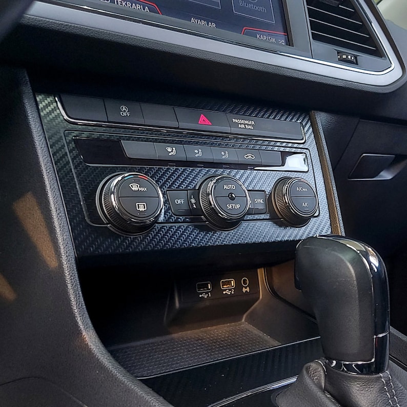 Puede incluir: Un salpicadero de coche negro con un patr&oacute;n de fibra de carbono. El salpicadero tiene varios botones, incluyendo "OFF", "AUTO", "SYNC" y "AC". Tambi&eacute;n hay dos puertos USB y una toma de auriculares.