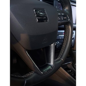 Puede incluir: Primer plano de un volante de cuero negro con un ribete de fibra de carbono y un detalle de costura blanca. El volante tiene el logotipo de SEAT y controles para el sistema de audio del coche.