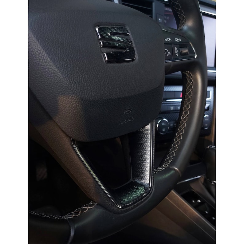 Puede incluir: Primer plano de un volante de cuero negro con un ribete de fibra de carbono y un detalle de costura blanca. El volante tiene el logotipo de SEAT y controles para el sistema de audio del coche.