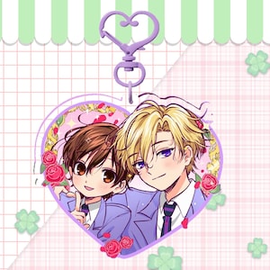 May include: A heart-shaped charm featuring two anime characters, framed by roses, with a lavender-colored heart-shaped clasp. The charm has a pink and white checkered background with green clovers.