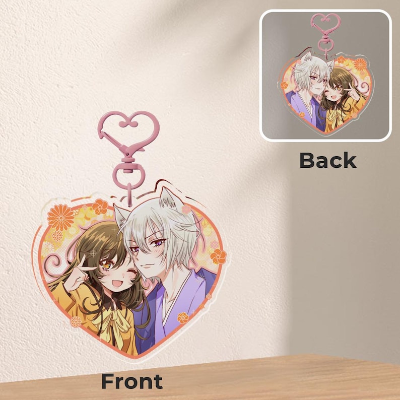 May include: Heart-shaped acrylic keychain featuring anime characters. The front shows a smiling girl and a man with white hair, while the back displays a similar image. The keychain has a pink heart-shaped clasp and a floral design.