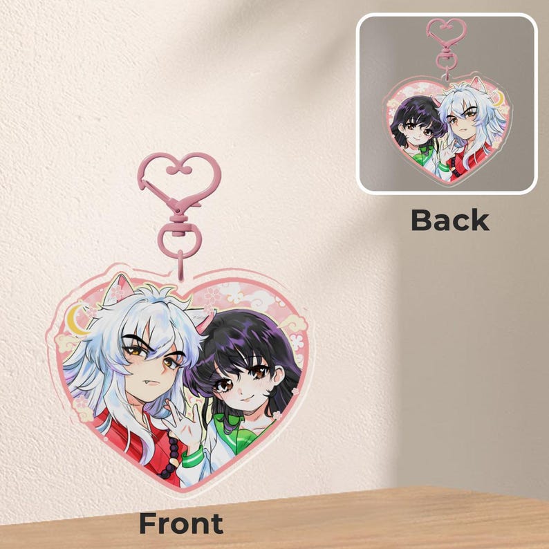 May include: A heart-shaped acrylic keychain featuring anime characters. The front shows two characters with white and black hair, wearing red and green outfits. The back of the keychain is also visible, with the same design. A pink heart-shaped clasp is attached.