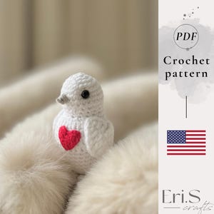 Op de afbeelding: Een handgemaakte witte gehaakte vogel met een rood hart op de borst. De vogel heeft een grijze snavel en zwarte ogen. De afbeelding bevat de tekst "Crochet pattern" en de Amerikaanse vlag. De Etsy shop naam "Eri.S crafts" is ook zichtbaar.