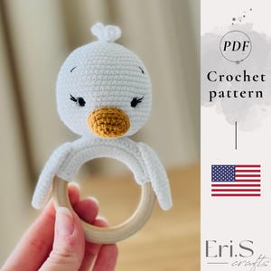 May include: A handmade white crochet duck rattle with a wooden ring. The duck has a yellow beak, black eyes, and a small tuft of white yarn on its head. The image also includes the text "Crochet pattern" and the American flag.