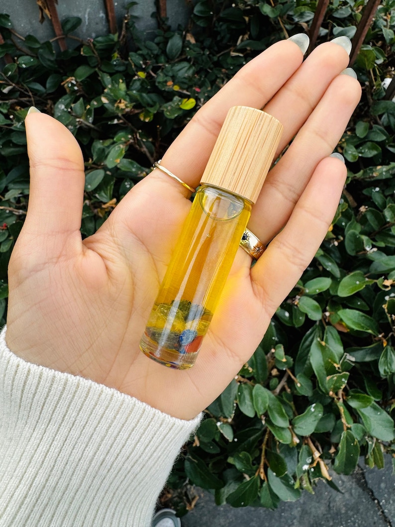 Pu&ograve; includere: Un flacone di vetro trasparente con rullo e tappo in bamb&ugrave;, riempito di un liquido giallo e cristalli colorati. Il flacone &egrave; tenuto in una mano.